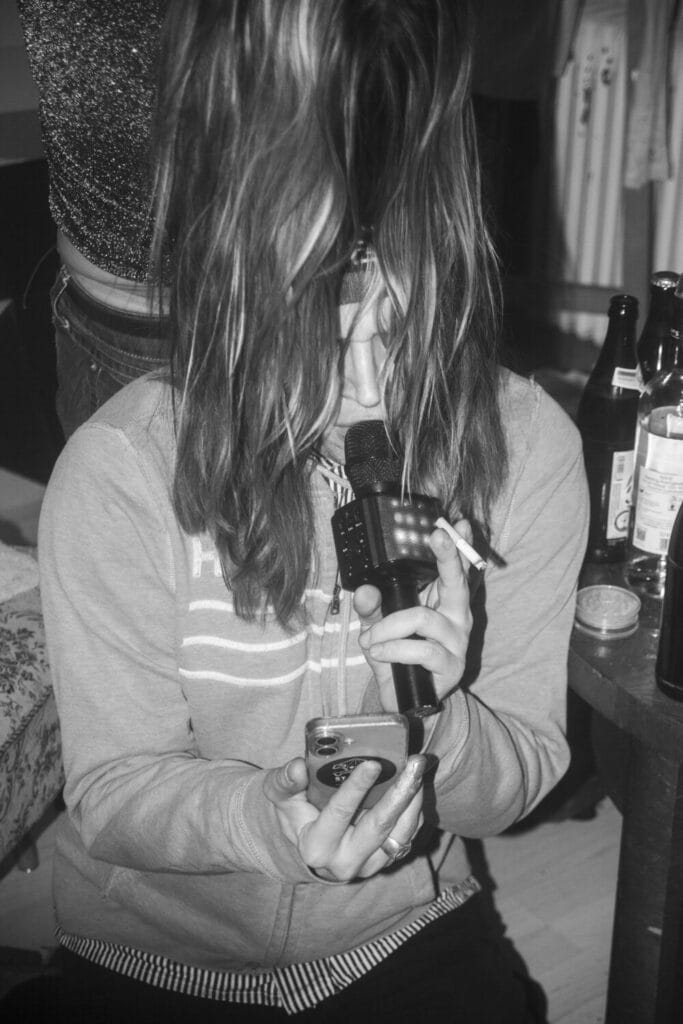 Erik, guy with karaoke mic and phone in hand and two wigs smoking and singing.