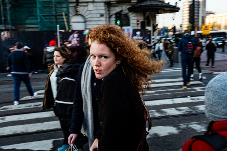 A woman with curly red hair and a serious expression looks over her shoulder while walking across a busy city crosswalk, captured by Stanley Robben, a self-taught street photographer from the Netherlands. Blurred pedestrians and urban buildings fill the background.