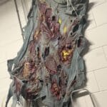 Ruth Spetter. The image depicts a piece of fabric or clothing that appears to be in a state of severe deterioration and decay. The material has a rotten, damaged texture and is dominated by grayish-purple and light blue tones. Its surface is characterized by numerous tears, depressions, and wrinkles, with scattered small red spots visible in certain areas. The background shows an indoor environment featuring metal chairs, highlighting the old and ruined condition of the fabric. There are no documents or tables present in the scene.