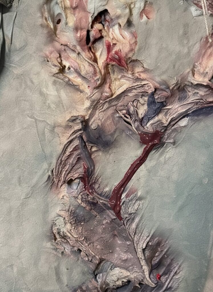 Ruth Spetter. The image depicts a piece of fabric or clothing that appears to be in a state of severe deterioration and decay. The material has a rotten, damaged texture and is dominated by grayish-purple and light blue tones. Its surface is characterized by numerous tears, depressions, and wrinkles, with scattered small red spots visible in certain areas. The background shows an indoor environment featuring metal chairs, highlighting the old and ruined condition of the fabric. There are no documents or tables present in the scene.